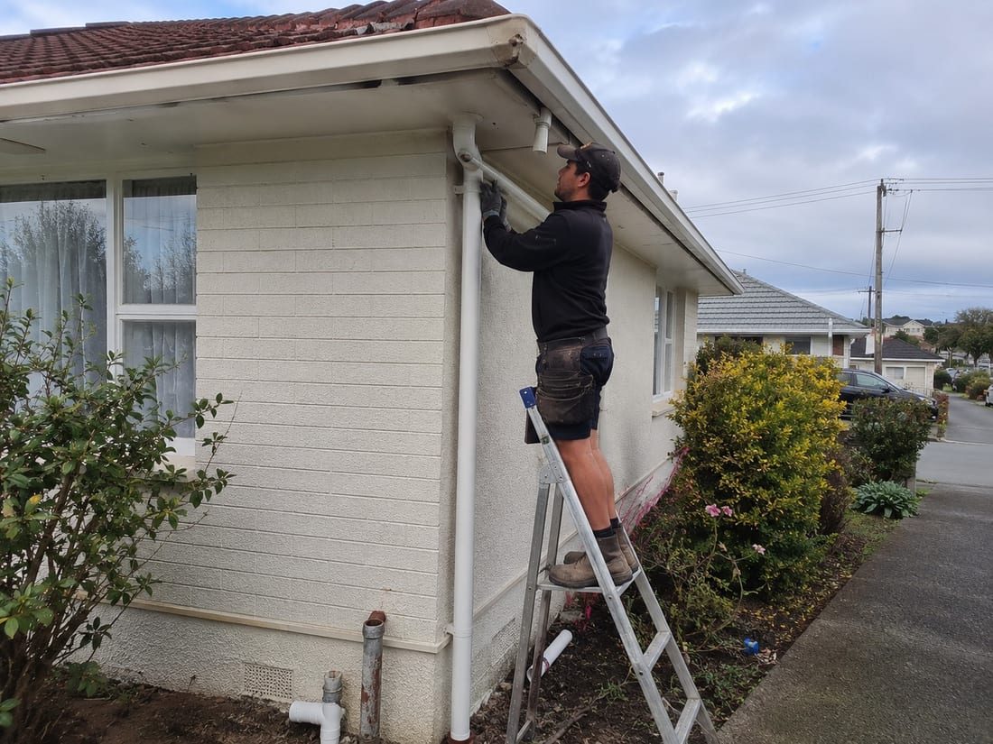 Installer on a ladder replacing the downpipe on the corner of an older Auckland home, with the old galvanised pipe removed at ground level
