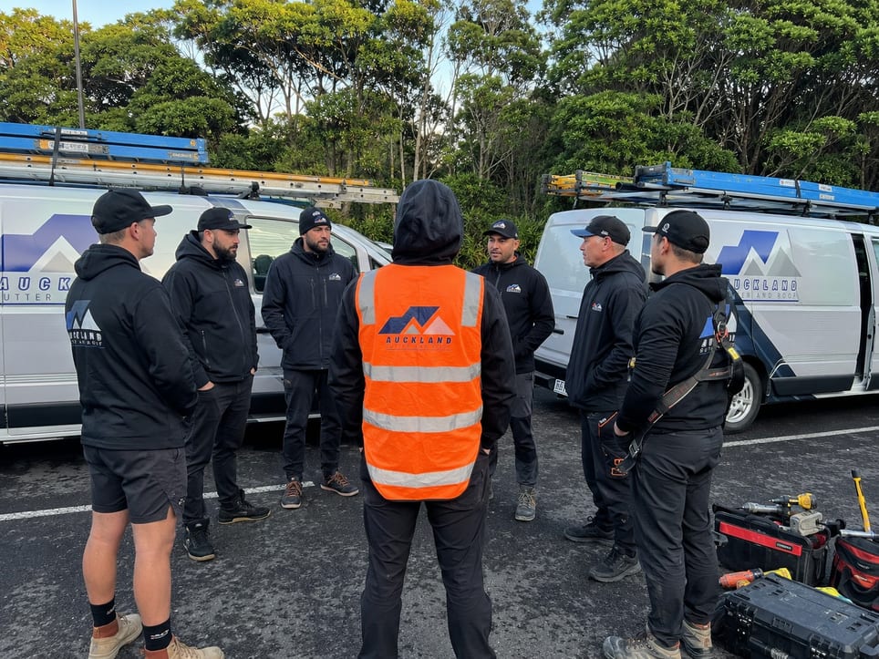 The Auckland Gutter and Roof team in branded workwear at a morning briefing beside their vans