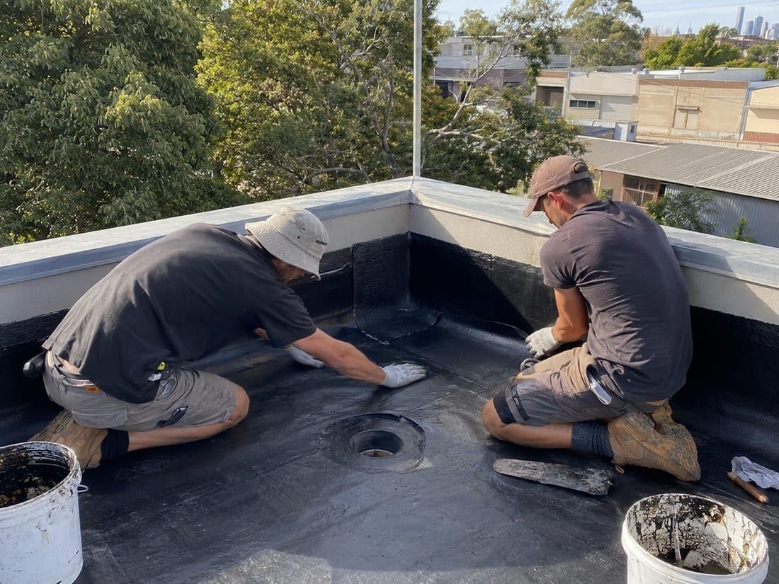 Two roofers sealing the membrane around a flat-roof drain outlet in Auckland